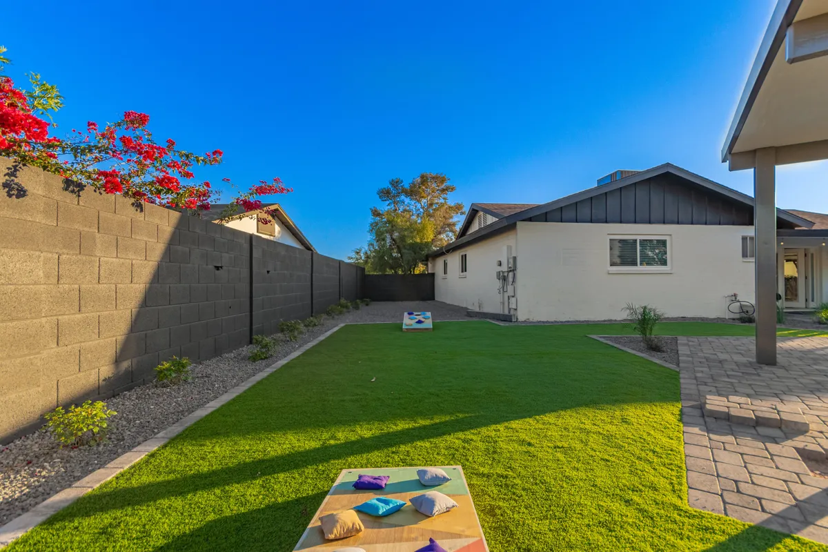 A cheerful backyard space with colorful bean bags scattered across a green lawn, adjacent to a paved patio. The setup is designed for outdoor games and informal social gatherings, encouraging friendly competition and playful interaction among guests.