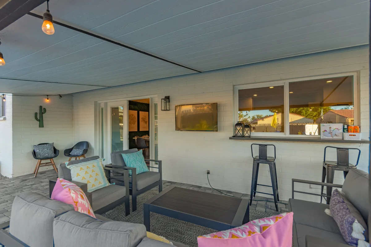 A comfortable outdoor seating area is arranged around a TV mounted on the wall, accented with colorful cushions and a decorative cactus. The space is inviting, perfect for watching sports, movies, or simply enjoying refreshments with friends.