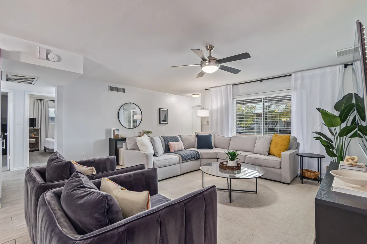 The photo displays a cozy sectional sofa, coffee table, decorative plants, wall art, and a mirrored wall decoration, creating a comfortable and welcoming setting. The focus is on the living room’s plush furnishings, ideal for socializing or relaxing with a movie night.
