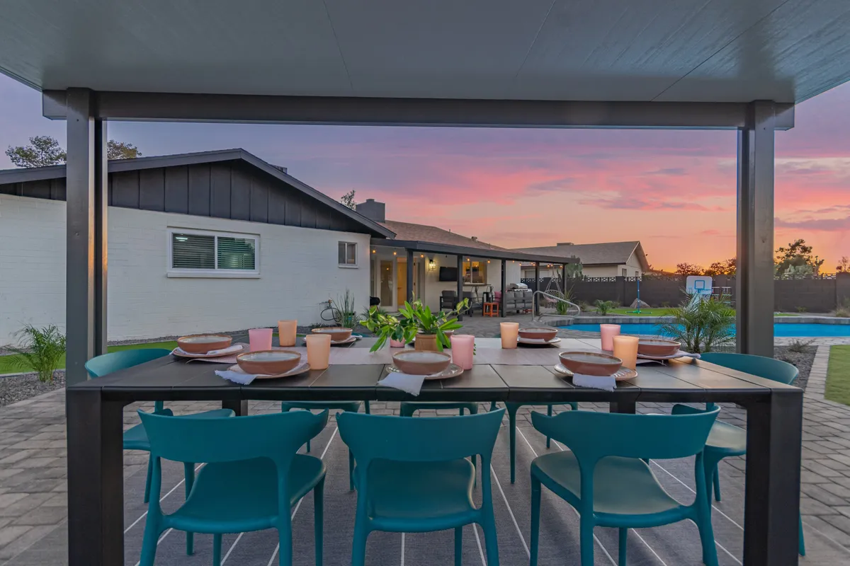 The scene features an outdoor dining table beside a sparkling pool under a vivid sunset sky, suggesting the perfect setting for social gatherings, relaxation, and unwinding after a day spent at nearby sports or entertainment venues.