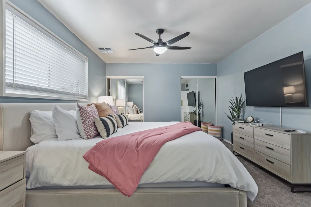 A cozy bedroom view showcasing a bed with decorative pillows, a TV atop a dresser, and vibrant plants, all set in a comfortable, relaxed atmosphere.