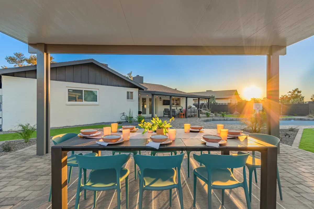 A vibrant exterior scene featuring a large outdoor dining table set up poolside, with a glowing sunset in the background. The pool sparkles invitingly, and there is ample space for guests to gather, eat, and relax. The atmosphere is lively and ideal for group gatherings, with the potential for both socializing and relaxing under the open sky.