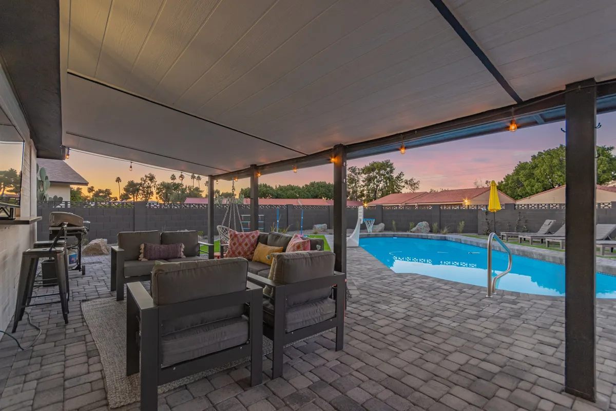 A vibrant backyard scene with a swimming pool, outdoor seating area under string lights, and a yellow umbrella. The area is ready for evening gatherings, poolside relaxation, or enjoying fresh air after logging off work.