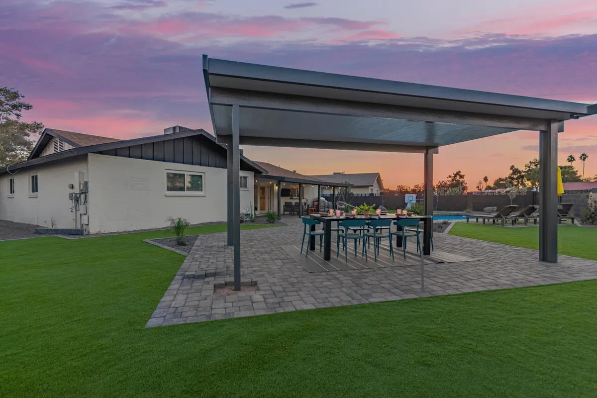 This image displays a welcoming outdoor dining area set on a landscaped lawn underneath a covered patio. The space is perfect for al fresco meals or work breaks in the fresh air, offering both sun and shade. Lush greenery and stylish patio furniture create a relaxed atmosphere ideal for unwinding between work sessions.