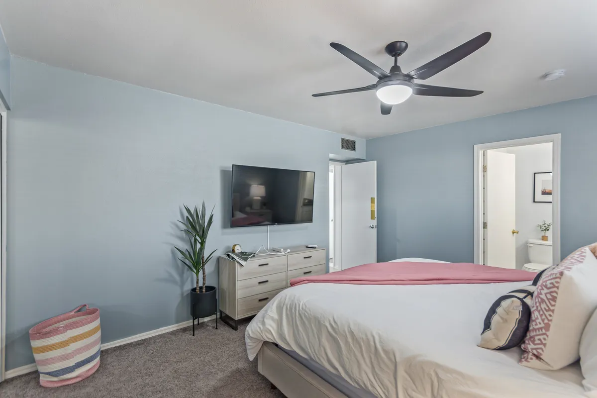 A tranquil bedroom with a bed dressed in a soft pink throw, a wall-mounted TV, a lush green plant in the corner, and a spacious dresser. The room receives natural light and has calming décor.