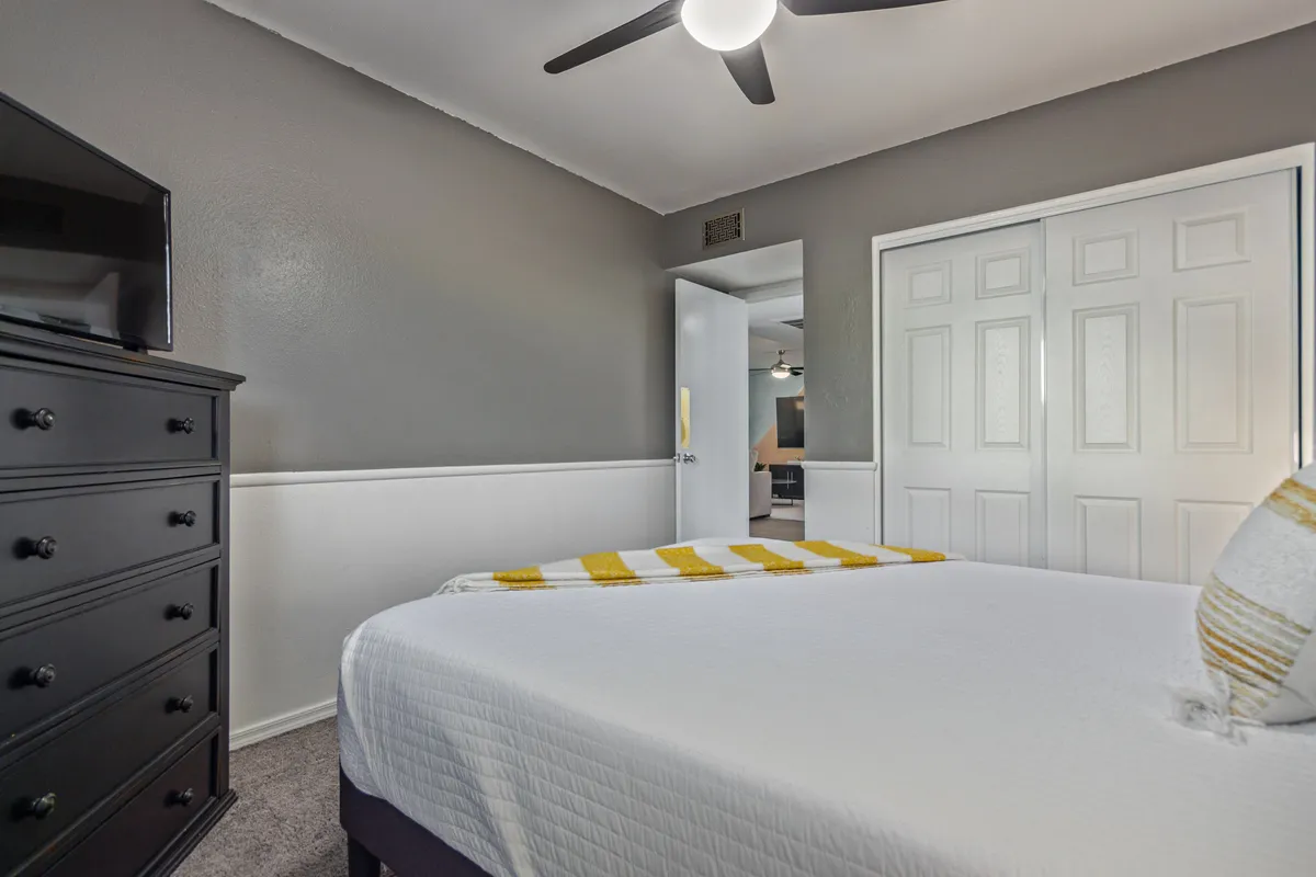 A cozy bedroom featuring a bed with a yellow striped blanket, a black dresser, and white closet doors. Soft daylight streams in, and a laptop is visible on the bed setup, suggesting both relaxation and work. The space is uncluttered, relaxed, and welcoming—ideal for productivity or downtime.