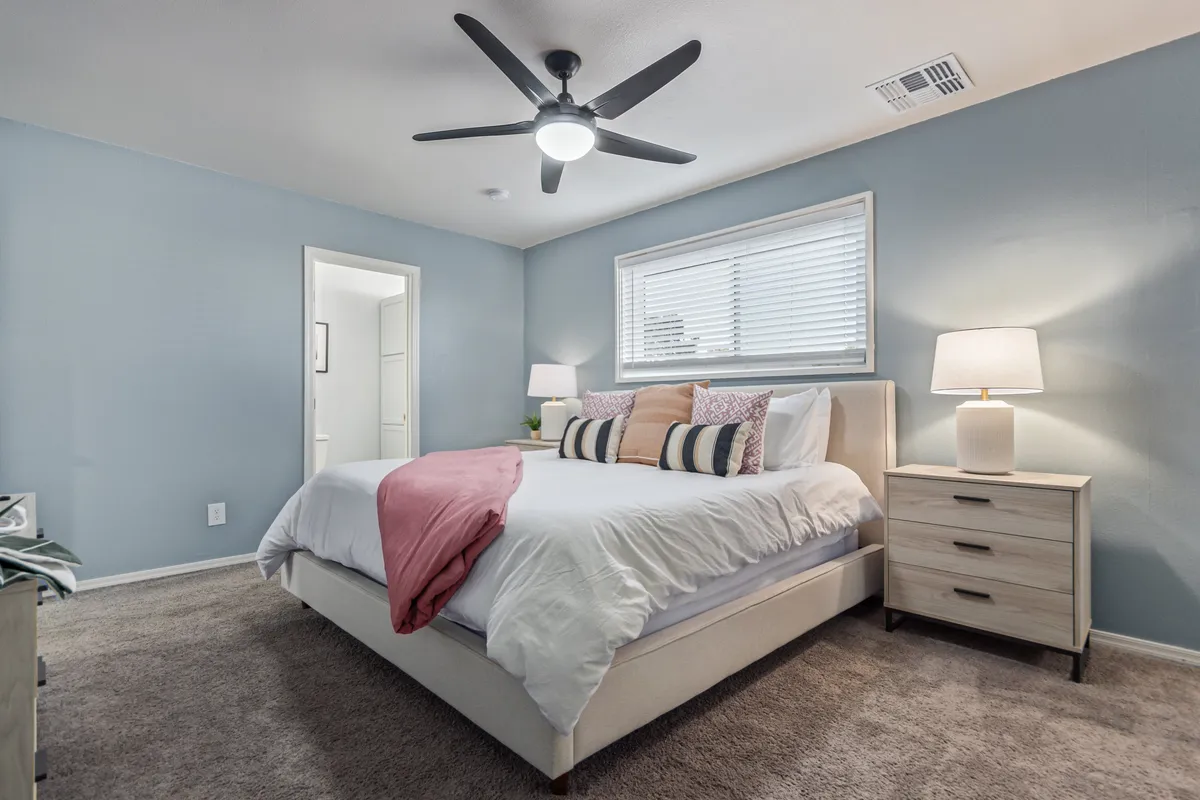 A tranquil bedroom scene with a restful bed adorned with decorative pillows, a nightstand with a glowing lamp, and a window covered by blackout blinds, creating a peaceful retreat.